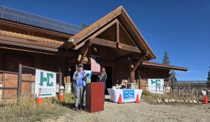 Two people stand at a podium outside Fairplay High Rockies Community School, a wooden building with solar panels. A table, shovels, and construction signs suggest a groundbreaking event. An American flag hangs above the entrance.