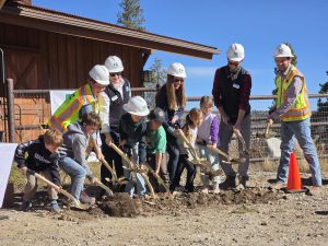 A group of adults and children from Fairplay High Rockies Community School, wearing hard hats and safety vests, use shovels to break ground at a construction site, smiling and posing in front of a wooden building.