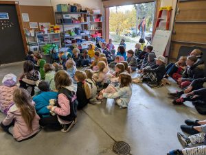 A group of children at High Rockies Community School sit on the floor of a garage-like room, facing forward. Shelves with supplies and a whiteboard are visible. The door opens to a sunny Fairplay outdoor area where two adults are standing.