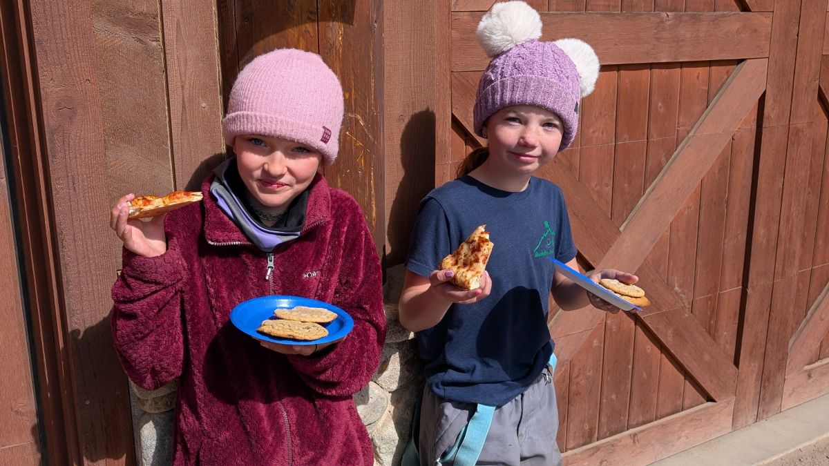 Two kids wearing knit hats and warm clothes hold blue plates with pizza and cookies while standing outside in front of a wooden wall, enjoying their food and smiling at the camera.