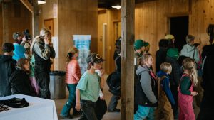 A group of children in casual clothing walk indoors through a wooden building, some looking forward and others glancing around. An adult stands nearby, and a table is visible in the foreground.