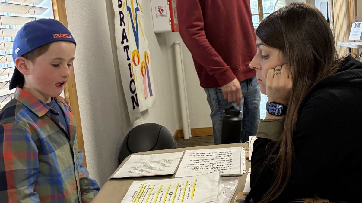 A young boy in a plaid shirt and blue cap speaks to a woman with long brown hair, who is looking at papers on a table. The woman appears to be listening attentively while resting her face on her hand.