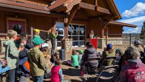 A group of people, including children and adults, sit outdoors in front of a wooden building, listening to a speaker at a podium. Some people are standing and clapping while others sit in chairs, enjoying the sunny day.