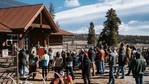 A group of people gathers outdoors in front of a wooden building with a red roof, listening to a speaker at a podium. Trees and open land are visible in the background under a partly cloudy sky.