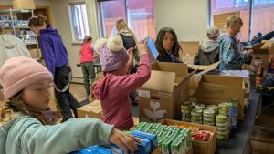 Children sort and organize food items such as canned goods and boxes in a room, packing them into cardboard boxes, likely as part of a food drive or community service activity.