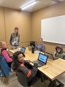 Four children sit at a table using laptops, smiling at the camera. An adult woman stands next to them, also smiling. They are in a classroom with tan walls and a large whiteboard in the background.