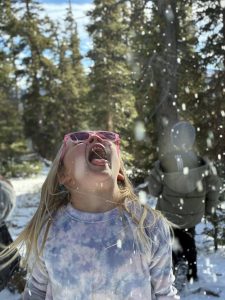 A young girl wearing pink glasses and a tie-dye shirt looks up joyfully with her mouth open to catch falling snowflakes, with snow-covered trees and another person in a winter coat in the background.