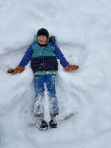 A child in winter clothes lies on their back in the snow, smiling while making a snow angel. Snow covers the ground and light flakes are falling.