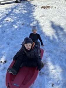 Two young children in winter clothes sit on a red sled on a snowy hill. One child is sitting on the sled while the other stands behind, both looking toward the camera. There are shadows and a parked car in the background.