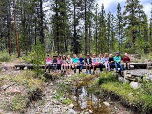 A group of children sit on a wooden bridge in a forest, smiling for the photo. Trees and greenery surround them, and a small stream runs under the bridge. The kids are dressed in colorful clothes.