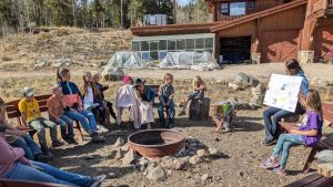 A group of children sits on benches around a fire pit outdoors, listening to a girl holding a poster. There is a log cabin and a greenhouse in the background, and the setting is rustic and surrounded by trees.