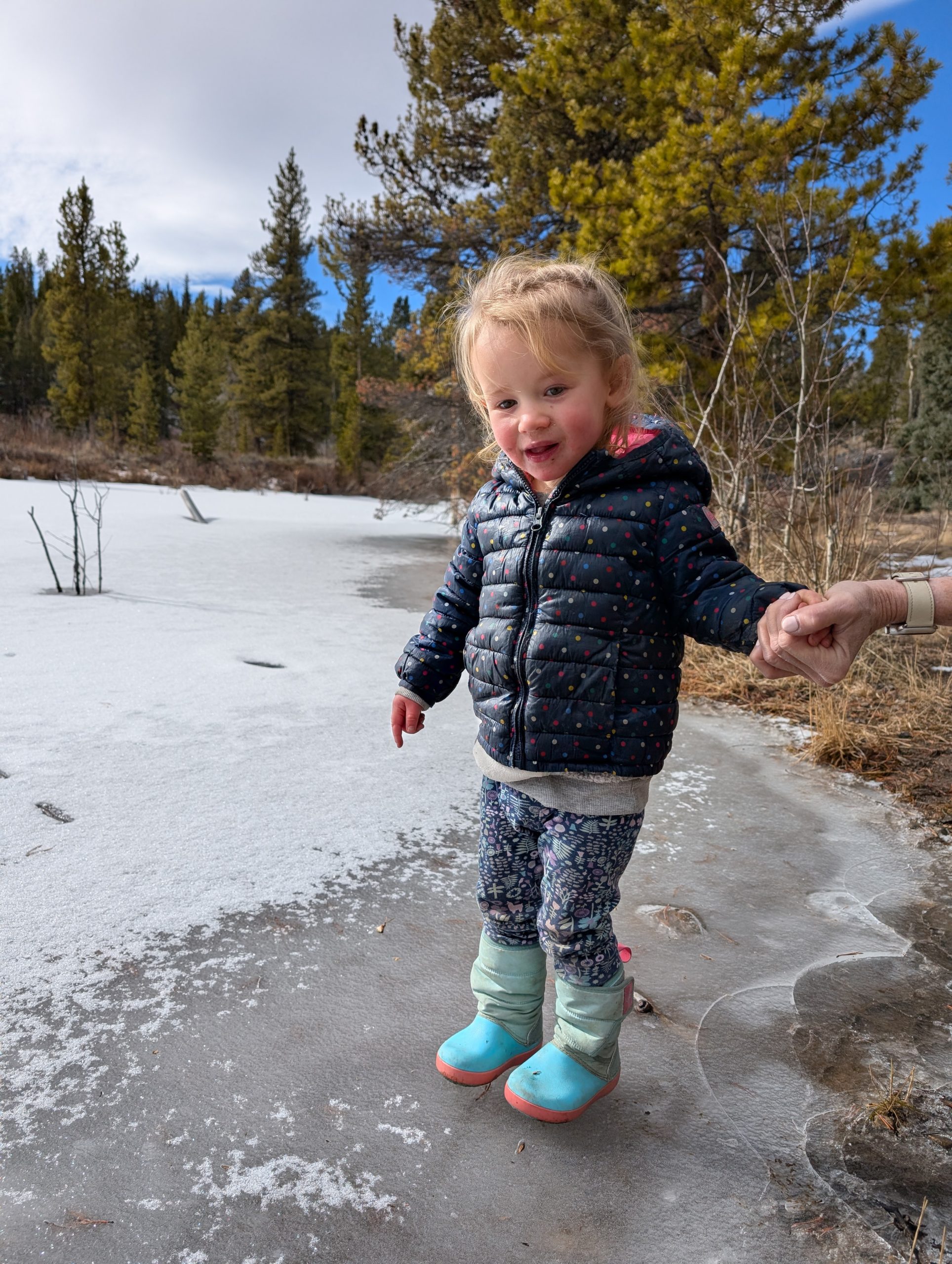A young child in a puffy jacket and boots smiles while standing on a frozen, snowy surface outdoors, holding an adults hand, with trees and a partly cloudy sky in the background.