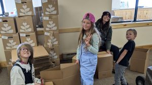 Four children are packing cardboard boxes in a room with stacked boxes covered in white leaf designs. One child flashes a peace sign, and everyone looks cheerful and engaged in their task.