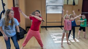 Five young girls are dancing and having fun in a well-lit indoor room with wooden floors and gym equipment in the background. They appear to be following dance moves, smiling and moving energetically.