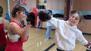 A woman wearing boxing gloves and pads smiles as a young girl in a white hoodie punches a pad in a gym room, both enjoying a boxing session.