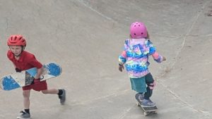 Two young children wearing helmets are at a skate park. One child in a red outfit carries a skateboard, while the other, in a colorful jacket and pink helmet, rides a skateboard on the concrete surface.