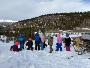 A group of children dressed in colorful winter clothes stand and sit in the snow, facing a snowy landscape with pine trees and mountains in the background. An old wagon with yellow wheels is on the right side.