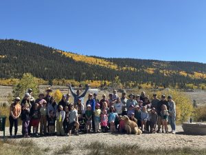 A large group of adults, children, and two dogs pose outdoors on a sunny day, with forested hills and golden autumn trees in the background. Everyone smiles and stands on a dirt area beneath a clear blue sky.