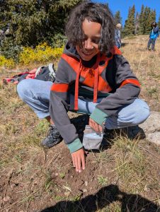 A child in a hoodie and jeans crouches outdoors, smiling while planting seeds in the soil. Pine trees, dry grass, and other people are visible in the background on a sunny day.