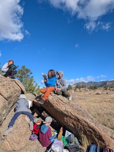 Several children sit and climb on large rocks outdoors, reading books under a bright blue sky with scattered clouds. Backpacks and other belongings are spread out on the rocks and ground nearby. Mountains are visible in the distance.