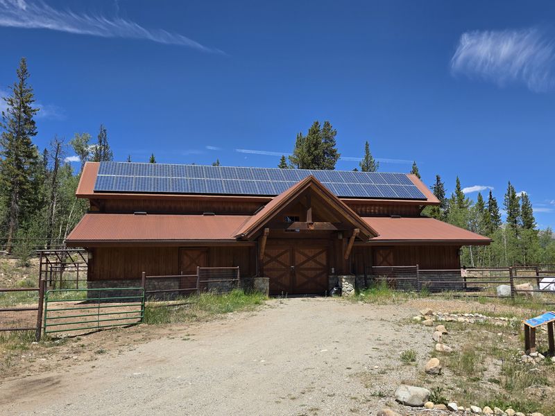 A wooden building with large solar panels on its roof is surrounded by a dirt path, fence, rocks, and trees under a clear blue sky.