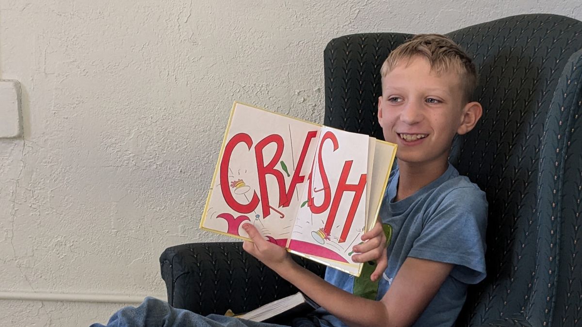 A smiling boy sits in a chair, holding up an open book that displays the word CRASH in large, colorful letters.