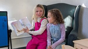 Two young girls sit together on a green armchair. One girl, wearing a bright pink outfit, holds up an open picture book and smiles at the camera, while the other girl in light blue overalls looks at her.