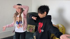 Two children stand indoors with arms raised, appearing to dance or pose. One wears a pink jacket and hat, the other a black hoodie. A chair, a large clock, and a book with a cartoon face are visible in the background.
