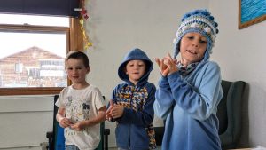 Three young boys stand indoors. Two wear blue hoodies; one also wears a blue knit hat with pom-poms. The third boy wears a white t-shirt. They appear to be clapping or rubbing their hands together.